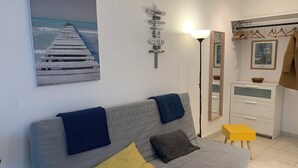 A modern living room with a grey sofa, a painting of a pier, a mirror, and a wooden coat rack. - Superb studio 2 to 4 people, seaside, casino and town center (Deauville)
