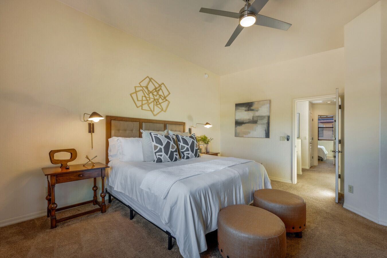 A bedroom with a bed, bedside tables, a desk, a ceiling fan, and a door leading to another room.