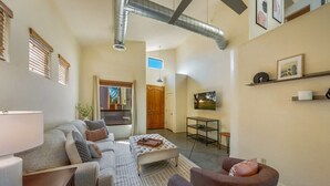 Ein geräumiges Wohnzimmer mit einer Couch, Sesseln und einem Couchtisch. Eine Tür führt in einen weiteren Raum, ein kleiner Tisch mit einer Lampe und Kunstwerke an den Wänden.
