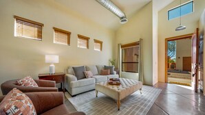 Ein geräumiges Wohnzimmer mit einer Couch, einem Sessel, einem Hocker, einem Couchtisch und einem großen Fenster mit Jalousien.