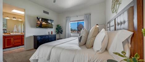 Un dormitorio con cama, mesita de noche, cómoda, ventilador de techo, ventana con cortinas y un cuadro en la pared.