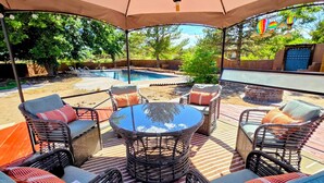 An outdoor patio area with wicker furniture and a round table, surrounded by a pool and trees.