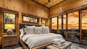 A wooden bedroom with a large bed, bedside tables, and a view of the sunset through large windows. - NEW! Heated Pool + Sauna + Dog-Friendly (Mineral Bluff)