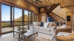 A spacious living area with a large window, a ceiling fan, and a staircase leading to an upper level. - NEW! Heated Pool + Sauna + Dog-Friendly (Mineral Bluff)