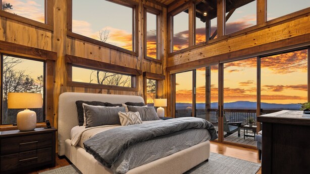 A bedroom with a large bed, wooden walls, and a view of the sunset through large windows. - NEW! Heated Pool + Sauna + Dog-Friendly (Mineral Bluff)