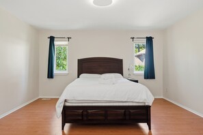 Une chambre à coucher avec un lit en bois, deux fenêtres avec des rideaux et une vue sur la verdure à l’extérieur.