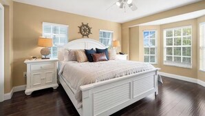 A bedroom with a large bed, two bedside tables, a ceiling fan, and a view of the outdoors through the windows. - Updated Key Largo Condo at Ocean Pointe - Private Beach & Balcony Views (Key Largo)
