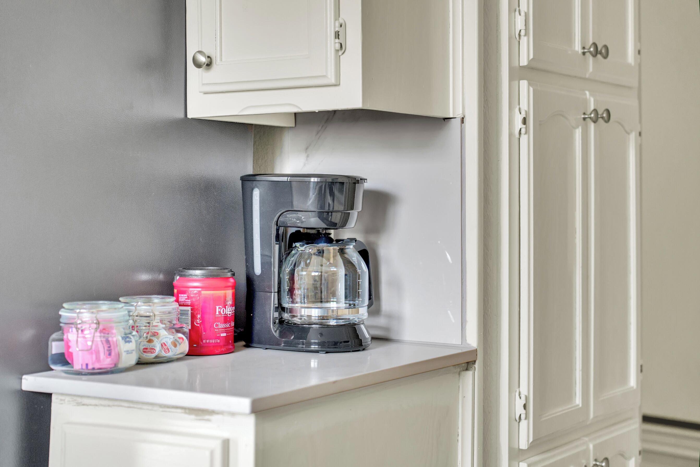 A kitchen counter with a coffee maker, jars of condiments, and a can of coffee.