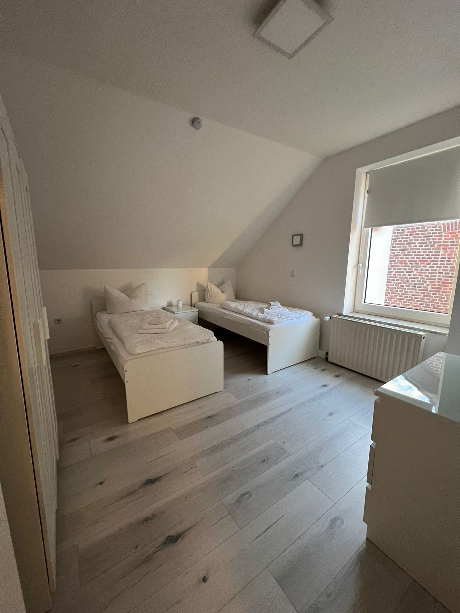 Een kleine, witte slaapkamer in loftstijl met een schuine kap, een raam met uitzicht op een bakstenen muur, en een radiator.