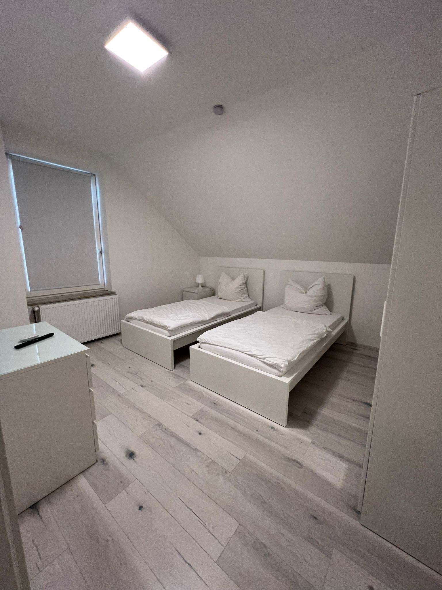 A bedroom with two single beds, a small bedside table with a lamp, a radiator, and a window with blinds.