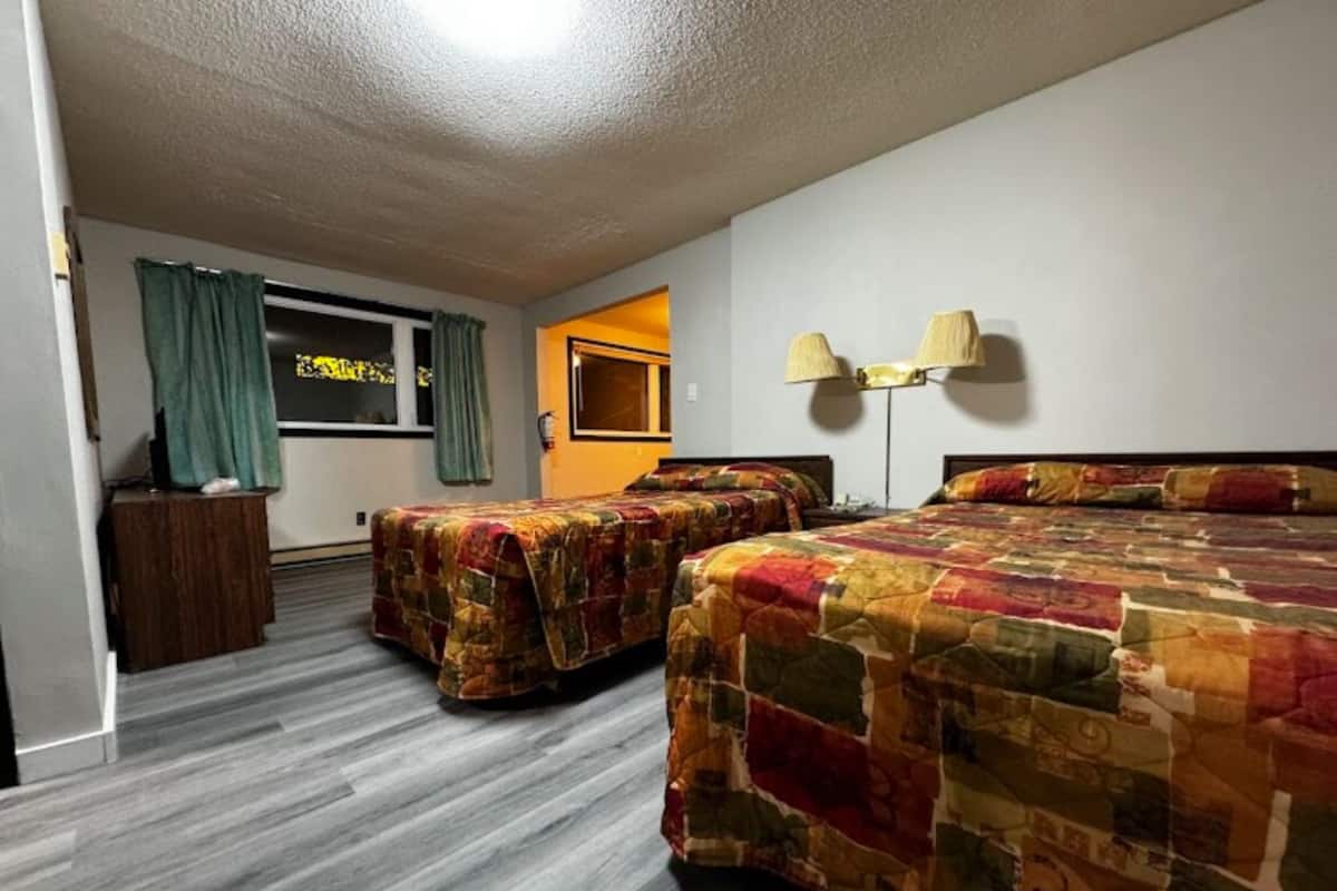 A hotel room with two beds, a wooden nightstand, a lamp, and a window with curtains.