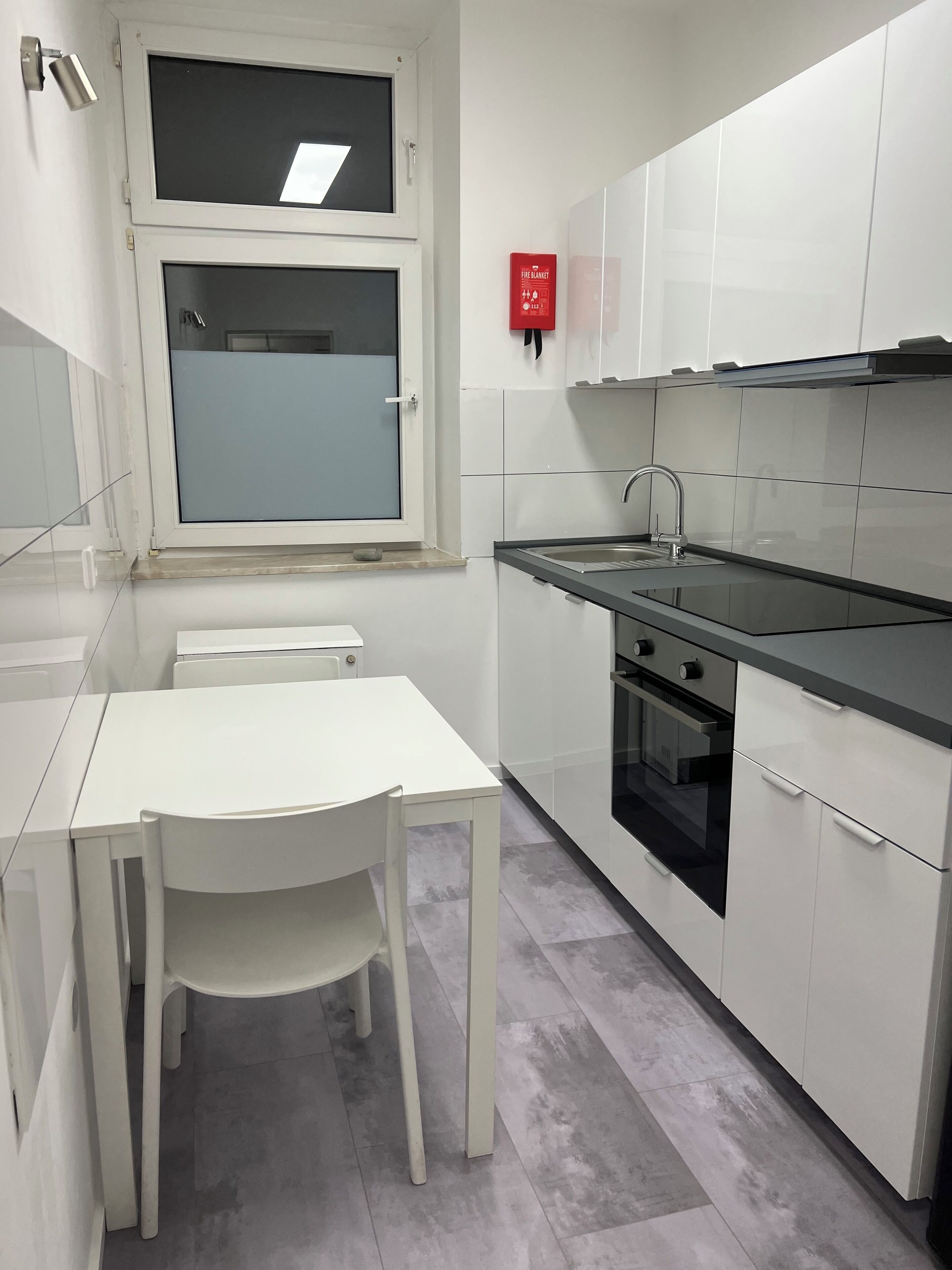 Eine moderne Küche mit weißen Schränken, einer schwarzen Arbeitsplatte, einem Spülbecken und einem Backofen. Ein Fenster bietet Blick ins Freie, und ein Feuerlöscher ist an der Wand befestigt.