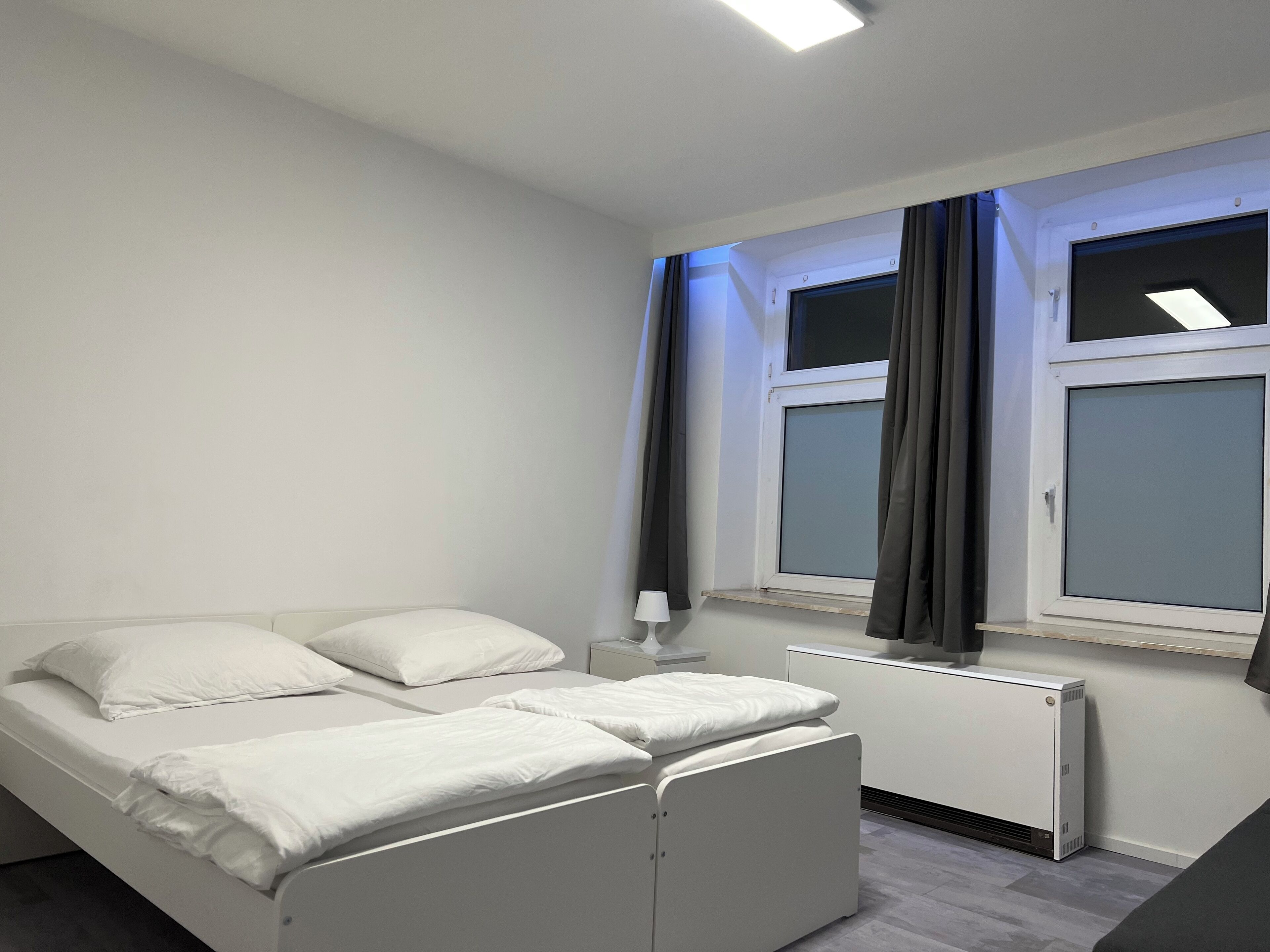 Une chambre à coucher avec un lit, une table de chevet, un radiateur et une fenêtre avec des rideaux.