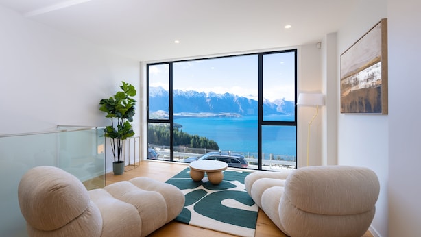 Un salon moderne doté d’une grande fenêtre offrant une vue sur les montagnes et un lac. On y trouve deux canapés beige, une table ronde et une plante en pot.