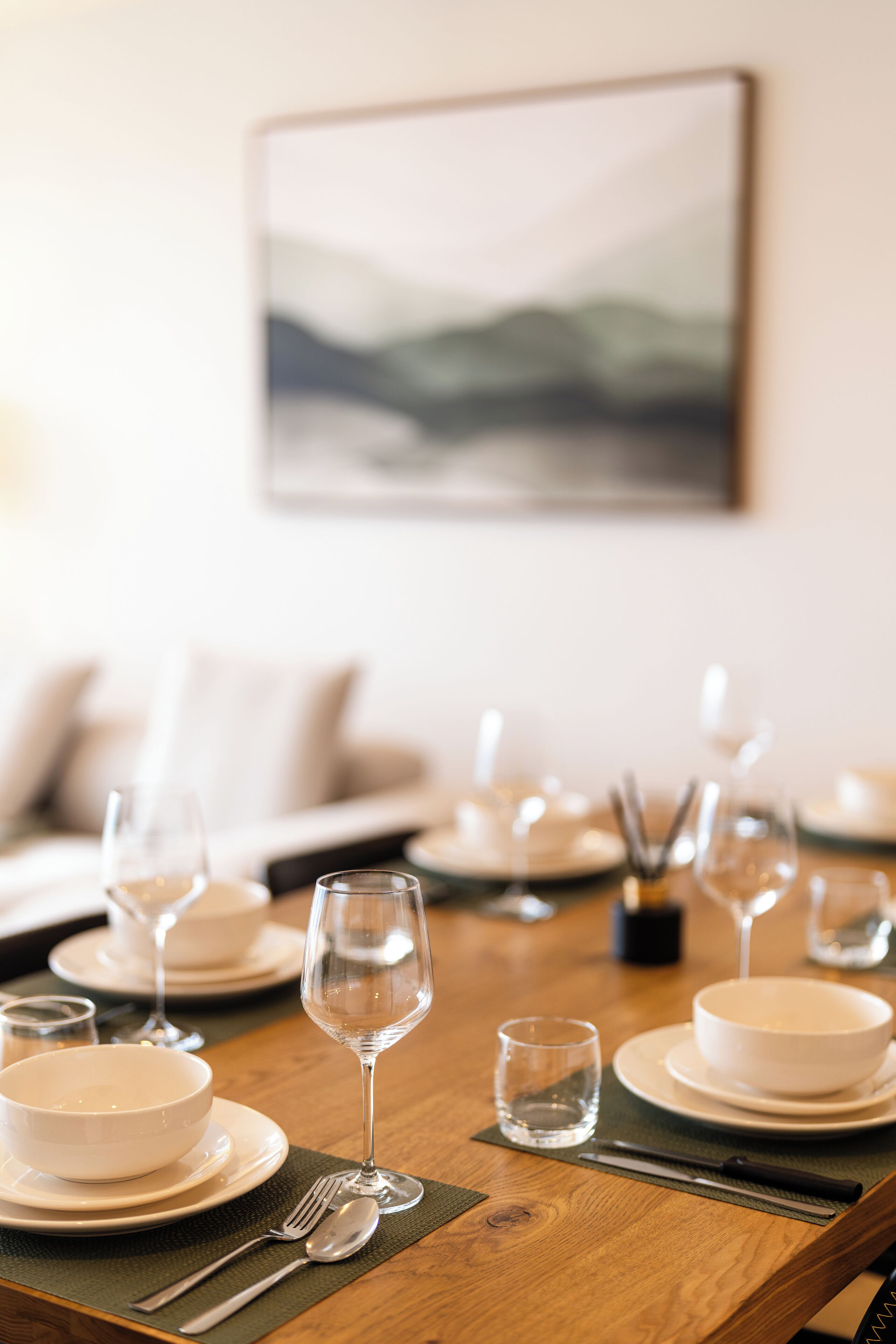 A dining table set with white plates, bowls, and wine glasses, with a painting on the wall behind.