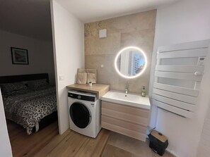 A compact hotel room with a washing machine, a sink with a mirror, and a bed with a leopard print cover. - Suite du garlaban jaccuzi (Aubagne)