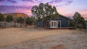 Un chalet rustique avec une terrasse en bois, entouré d’arbres et posé sur un sol en terre battue, le tout se détachant sur le ciel au coucher du soleil.