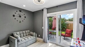 A modern living room with a grey sofa, a wall clock, and a sliding glass door leading to an outdoor patio. - Staycation Station (Tampa)