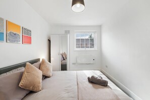 Une chambre à coucher avec un lit, des oreillers, une table de chevet, une fenêtre avec des stores et des œuvres d’art accrochées au mur.