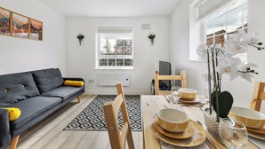 A modern living room with a grey sofa, wooden dining table set with yellow and white bowls, and a large window with a view of buildings. - 4 Min to LTN | Modern Stylish | WiFi | FreeParking (Luton)