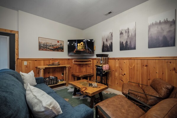 Un salon chaleureux avec un lambris en bois, un téléviseur à écran plat, un jeu d’échecs, un canapé en cuir marron et un divan bleu.
