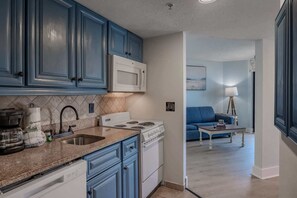 A kitchen with blue cabinets, a white microwave, and a stove. There is a sink with a faucet, a countertop, and a dishwashing machine. A blue sofa and a small table with a lamp are visible in the adjacent living area.