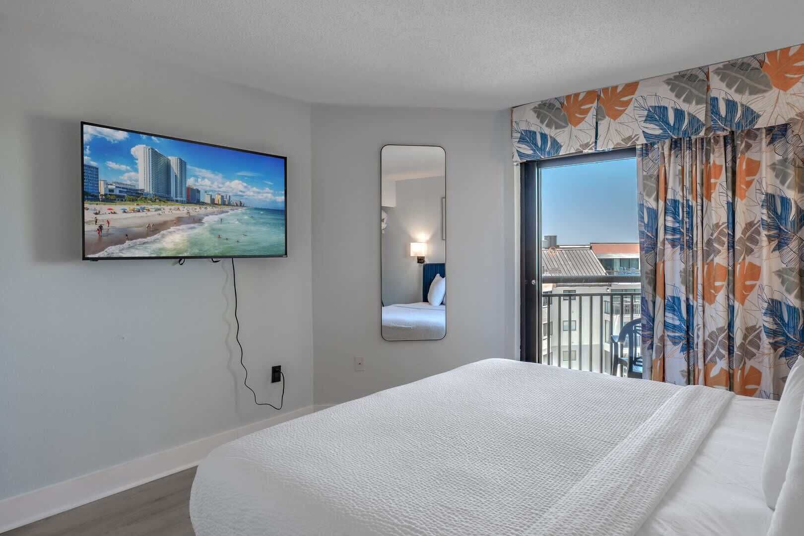 A hotel room with a bed, a TV mounted on the wall, a mirror, and a balcony with curtains.