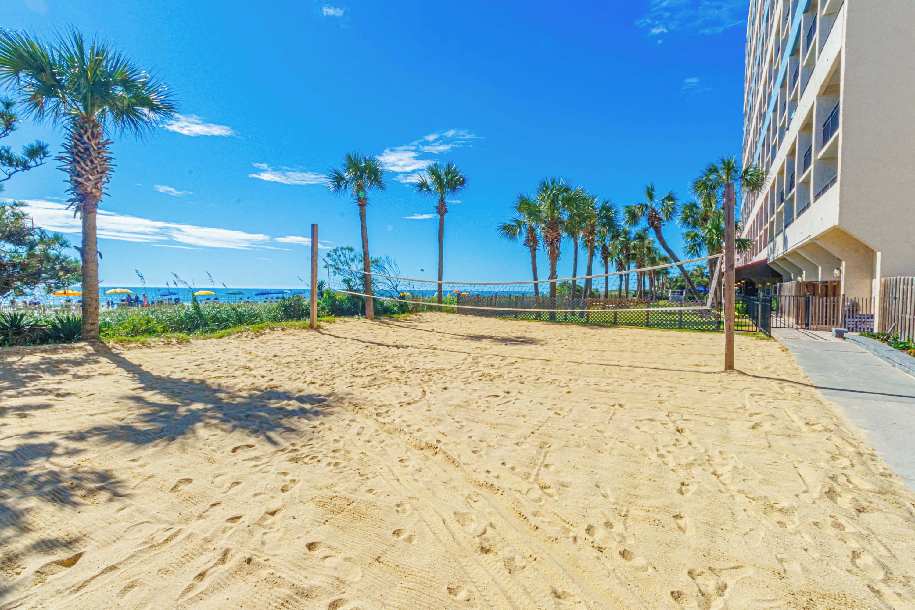 Un terrain de beach-volley sablonneux, avec des palmiers et un bâtiment en arrière-plan.