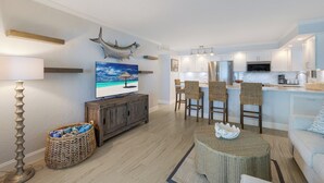 A modern living room with a TV, wicker furniture, and a kitchen area with white cabinets and stainless steel appliances. - BG Beach Club 3C | 3BR/2BA Boca Grande | Pool View (Boca Grande)