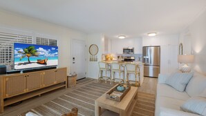 Un salon moderne avec un téléviseur, un coin repas avec des chaises, une cuisine équipée et un canapé blanc.