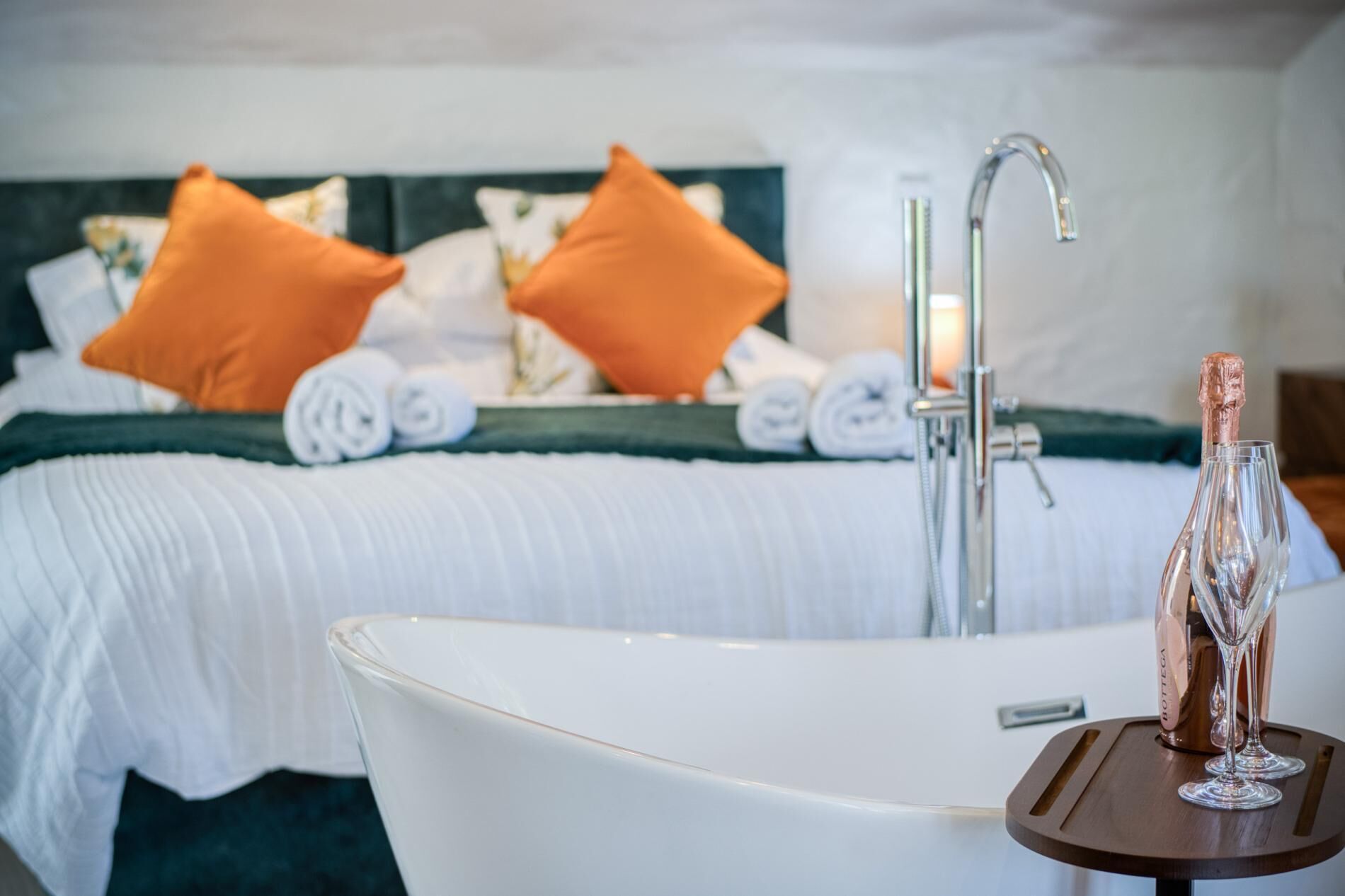 Une chambre d’hôtel avec une grande baignoire blanche, une bouteille de champagne et un lit parfaitement fait, agrémenté d’oreillers orange et de serviettes roulées.