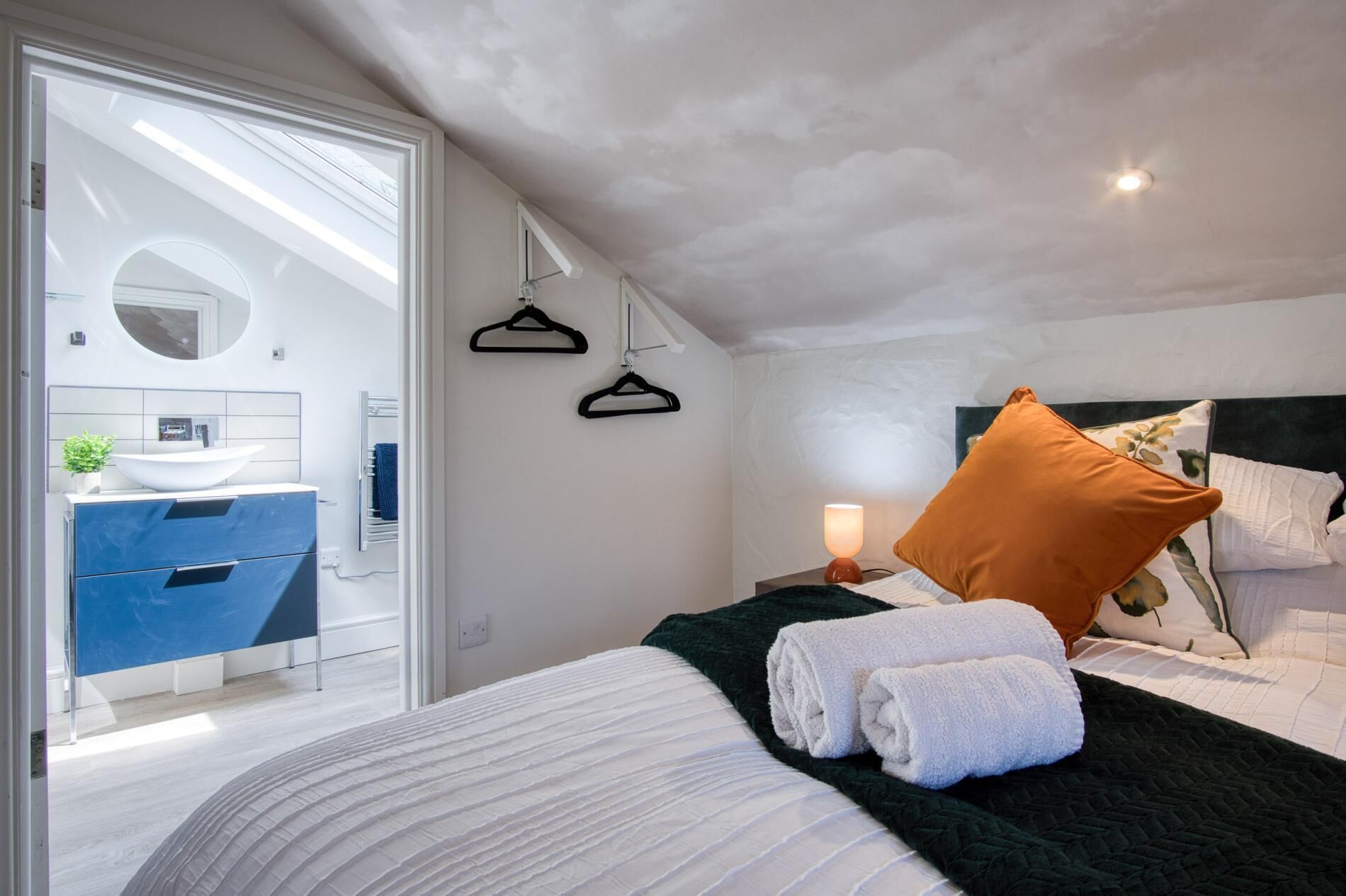 A modern bedroom with a bed, a bedside table with a lamp, a hanging light fixture, and a view of a bathroom with a sink and mirror.