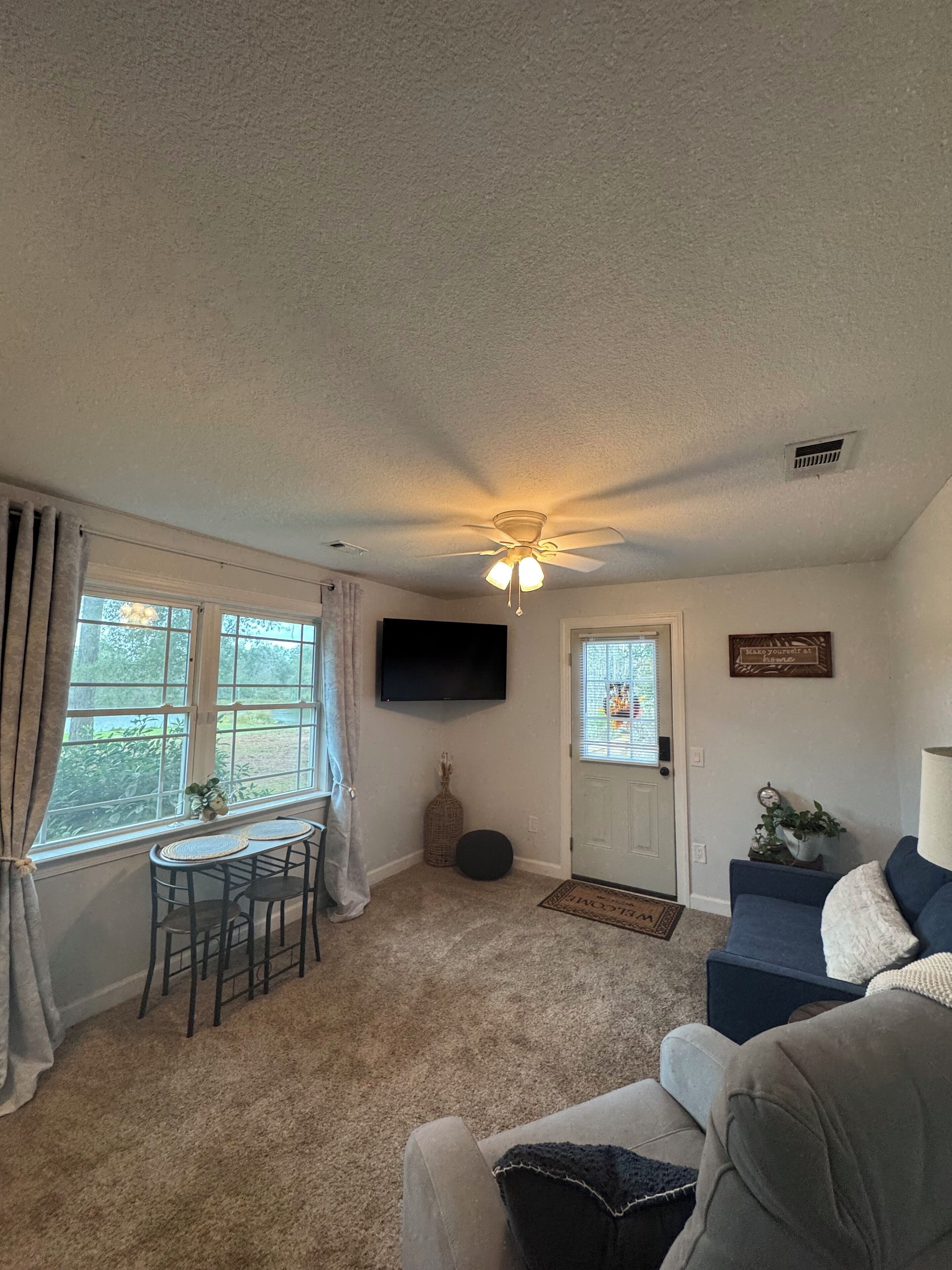 Uma sala de estar com ventilador de teto, televisão fixada na parede, área de refeições com mesa e cadeiras, e uma porta com placa decorativa.