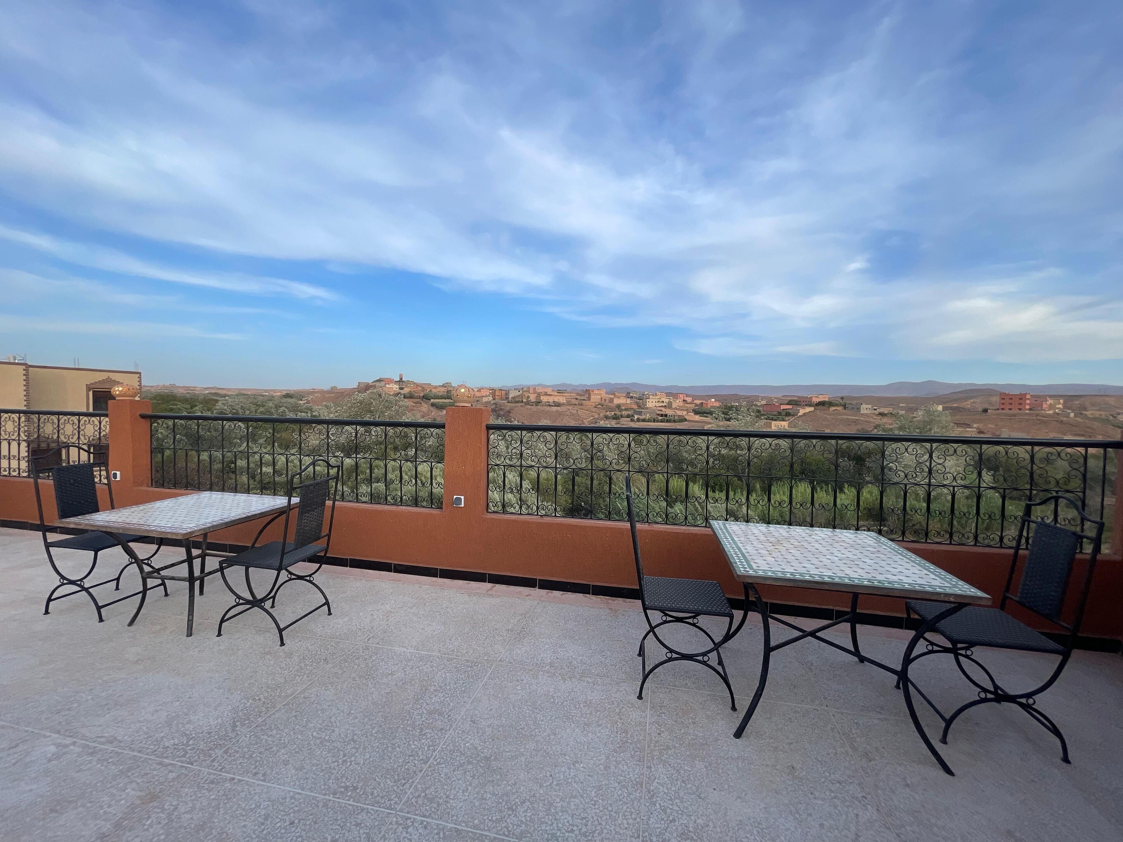 Eine Terrasse mit zwei Tischen und Stühlen, einem Metallgeländer und Blick auf entfernte Gebäude und Hügel.