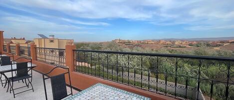 Una terraza con una mesa y sillas de cerámica, baranda de metal y vistas a un pueblo lejano y colinas.