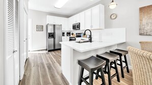 Una cocina moderna con gabinetes blancos, una isla central con taburetes, electrodomésticos de acero inoxidable e iluminación colgante.