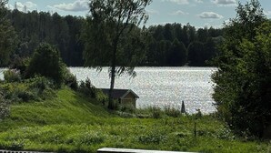 A lakeside view with a wooden bench in the foreground, a small shed, and a forested hill in the background. - Stylish Lakehouse retreat with views over Djulö (Katrineholm)