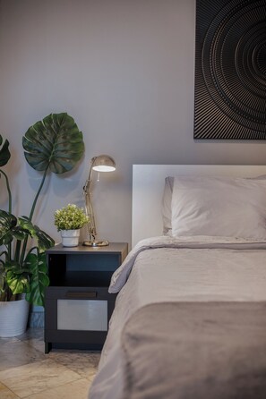 A neatly made bed with a white headboard, a bedside table with a lamp and a small plant, and a large green leaf decoration on the wall.