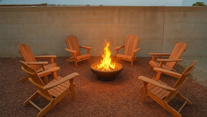 A fire pit with four wooden Adirondack chairs arranged around it, set on a mulch-covered ground with a brick wall in the background. - The Ultimate OC Stay – NEW Stylish 6BR Home Perfect for Families & Groups! (Santa Ana)