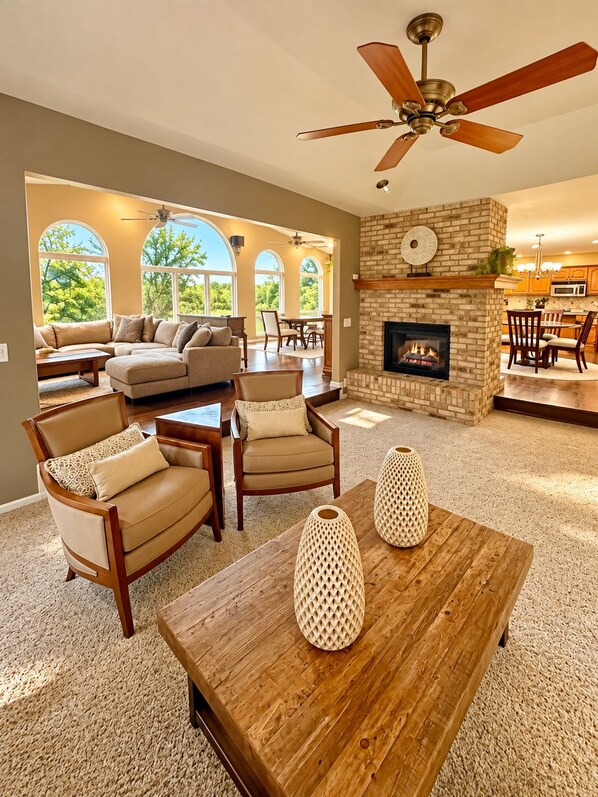 A spacious living room with a fireplace, a ceiling fan, and a view of the outdoors through large windows.