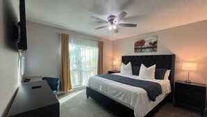 Habitación de hotel con cama, escritorio, ventilador de techo, televisión y ventana con cortinas.