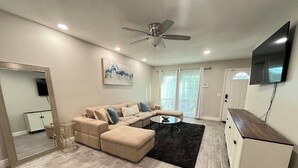Una sala moderna con un sofá seccional color beige, una mesa de centro de vidrio y un ventilador de techo.