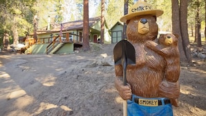 A wooden bear statue holding a shovel and wearing a hat labeled "SMOKEY" in a forested area with a cabin in the background. - One acre with a fire pit, kids play area, treehouse, zipline, pets welcome (Springville)