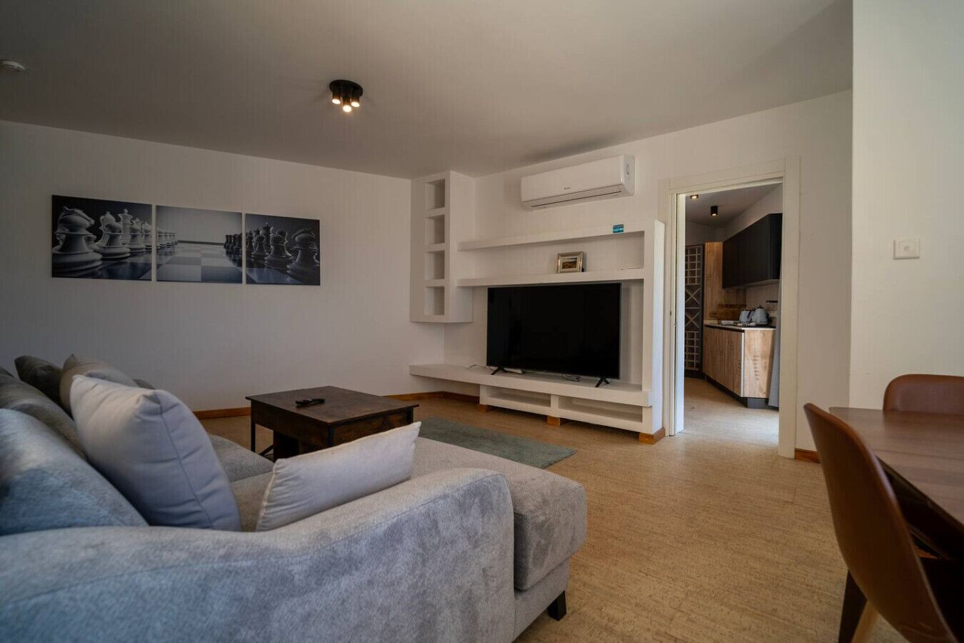 A modern living room with a grey sofa, a flat-screen TV, and a small table. There is a kitchen area visible through an open doorway.