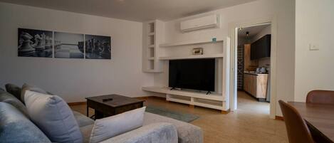 A modern living room with a grey sofa, a flat-screen TV, and a small table. There is a kitchen area visible through an open doorway.
