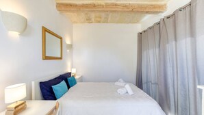 A hotel room with a large bed, a wooden headboard, a bedside table with a lamp, a mirror, and a window with curtains. - Authentic Charming Farmhouse With Pool in Gozo (Zebbug)