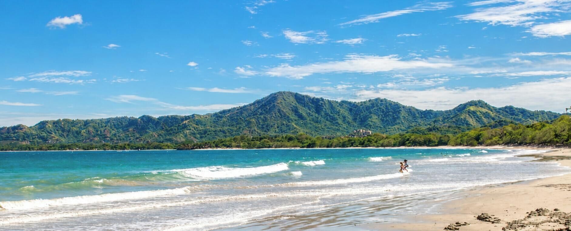 Una playa con agua cristalina, una persona corriendo y una montaña al fondo.