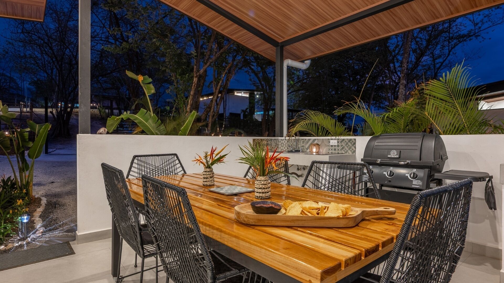 Une terrasse extérieure avec une table en bois, des chaises en osier noir, un barbecue et des plantes en pot.