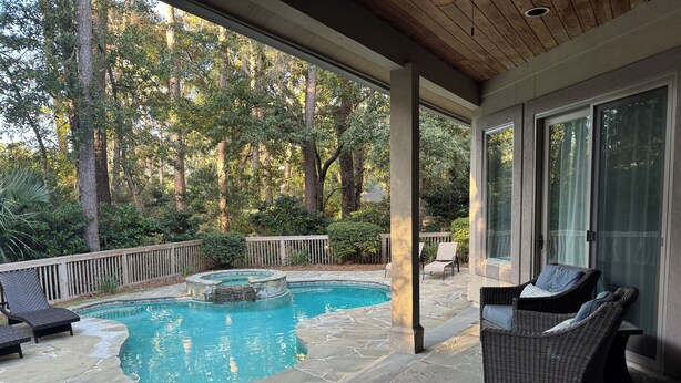 A covered outdoor swimming pool with stone flooring, surrounded by wooden deck chairs and a wicker lounge chair. - WALK TO BEACH! Pool/spa fenced yard. (Hilton Head Island)