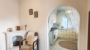 Ein Zimmer mit Kamin, einem Sessel und Blick in eine Küche durch einen Bogen.